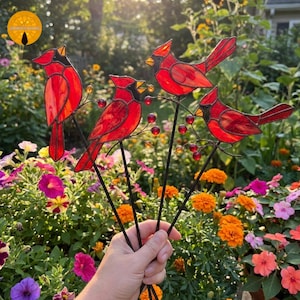May include: Four stained glass cardinal ornaments in vibrant red with black facial details, each perched on a thin metal stem adorned with small red glass berries. The birds feature translucent red glass panels held together by dark metal solder lines.