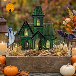 Op de afbeelding: Een spookhuis van glas-in-lood in groentinten met een zwart metalen frame, voorzien van spitsboogvensters en een puntdak. Het huis is omringd door een witte spookfiguur, een paarse vleermuis en kleine lichtjes in een houten kist.