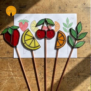 May include: Five stained glass garden stakes in the shape of fruit and leaves. The stakes include a red strawberry, a yellow lemon slice, two red cherries, an orange slice, and green leaves. Each stake has a copper stem and is set against a drawing.
