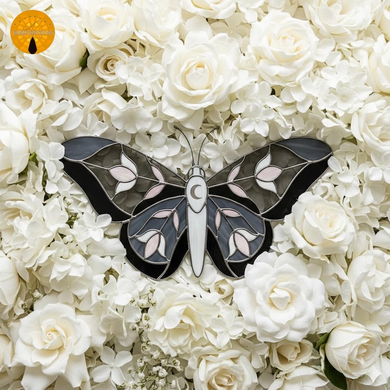 May include: A decorative butterfly with a stained glass design, featuring black, gray, white, and pink accents. The butterfly has a crescent moon detail and is set against a backdrop of white roses.
