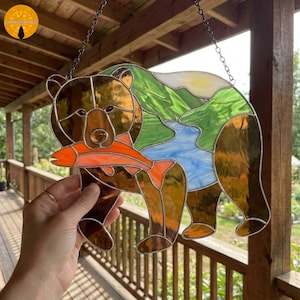 Könnte beinhalten: Buntglas-Kunstwerk eines Bären mit einem Lachs. Der Bär ist braun mit bernsteinfarbenen Akzenten, der Lachs orange, und der Hintergrund zeigt einen Fluss und Berge in Grün und Blau. Aufgehängt an einer Kette.