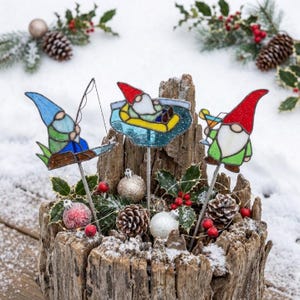 Puede incluir: Tres estacas de jardín de vidrio tintado con gnomos. Un gnomo pesca con sombrero azul, otro flota en una balsa amarilla con sombrero rojo y otro sostiene un cóctel con sombrero rojo. Cada gnomo está unido a una varilla de metal delgada.