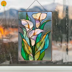 May include: Stained glass panel featuring calla lilies in shades of white, yellow, and purple, with green leaves. The artwork is framed in a copper-colored metal and hangs from a chain. The background is blurred, suggesting a window.