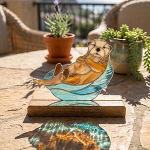 Pode incluir: Estatueta de lontra em vitral, repousando sobre uma onda azul, montada numa base de madeira. A lontra tem tons castanhos e bege, com um rosto claro. A estatueta é exibida ao ar livre com plantas ao fundo.