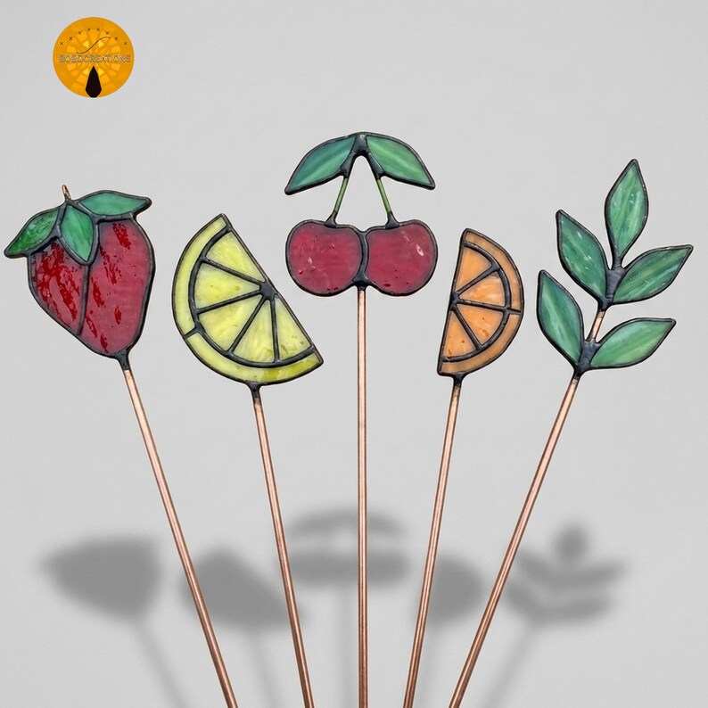 May include: A collection of five decorative garden stakes featuring colorful stained glass fruit and leaf designs. The stakes include a red strawberry, a yellow lemon slice, red cherries, an orange slice, and green leaves, all with copper stems.