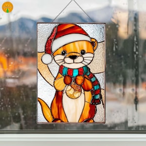 Pode incluir: Painel de vitral com uma lontra de desenho animado usando um gorro de Papai Noel e um cachecol listrado, segurando um enfeite dourado. O painel retangular é pendurado por uma corrente, perfeito para decoração de festas.