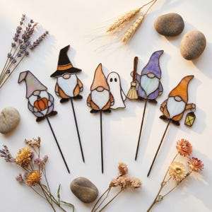 May include: Five stained glass garden stakes featuring gnomes with various accessories including a pumpkin, a witch hat, a ghost, a broom, and a lantern. Each gnome is crafted with colorful glass pieces and thin metal borders, attached to a thin metal rod.
