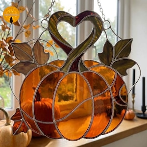 May include: A stained glass pumpkin suncatcher featuring orange and amber glass segments with dark green leaf accents. The top of the pumpkin stem forms a heart shape. The piece is framed in silver metal solder and hangs from a silver chain.
