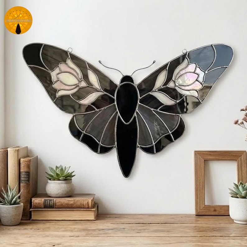 May include: A stained glass butterfly with a black body and wings in shades of gray, white, and pale pink. The wings feature floral designs and are outlined with a silver metal. The butterfly is hanging on a white wall.