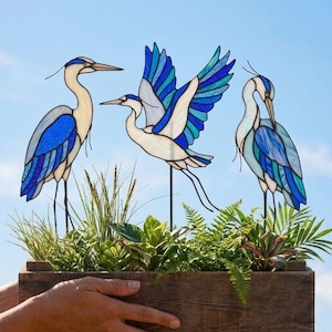 May include: Three stained glass heron figures in shades of blue, white, and teal, mounted on thin metal stakes. The herons are depicted in different poses, including standing and flying, with intricate glass paneling and dark metal soldering.