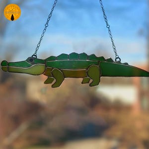 Op de afbeelding: Een gebrandschilderd glazen alligator zonnevanger in groen- en geeltinten, hangend aan een ketting. De alligator heeft een gedetailleerd ontwerp met een gesegmenteerd lichaam en poten. De zonnevanger is ontworpen om in een raam of aan een muur te hangen.