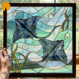 Puede incluir: Panel de vidrieras con dos rayas nadando en una escena submarina. La obra de arte utiliza tonos de azul, verde y vidrio transparente para representar el agua, las burbujas y las plantas acuáticas. El panel está enmarcado en un marco de madera marrón oscuro.