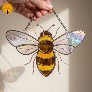 May include: A stained glass bee suncatcher with iridescent wings, a yellow and brown body, and copper wire details. The bee is suspended by a chain. A decorative piece for windows or walls.
