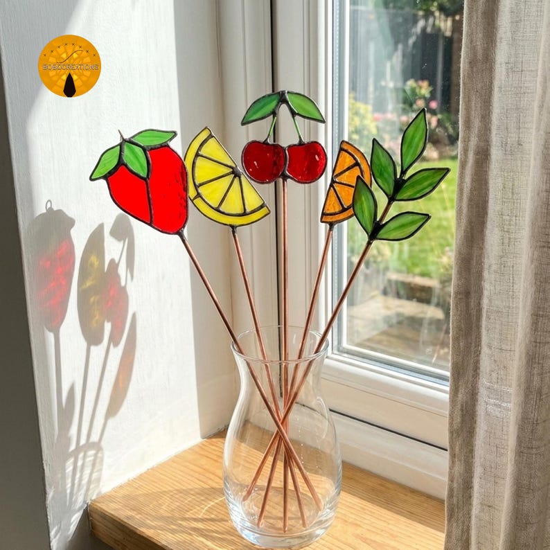 May include: A clear glass vase holds five colorful stained glass fruit garden stakes. The stakes feature a red strawberry, a yellow lemon slice, two red cherries, an orange slice, and green leaves. The stakes are copper-colored and are displayed near a window.