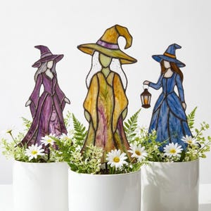 Könnte beinhalten: Drei Buntglas-Hexenfiguren in weißen Keramiktöpfen mit grünem Blattwerk und weißen Blumen. Die linke Hexe trägt ein violettes Gewand, die mittlere Hexe ein gelb-grünes Gewand mit hohem Hut und die rechte Hexe ein blaues Gewand mit einer Laterne.