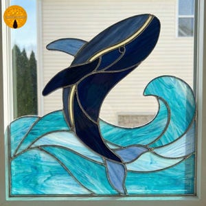 May include: Stained glass window panel featuring a breaching whale. The whale is depicted in shades of blue, with a cream-colored accent, against a backdrop of turquoise waves. The panel is framed in a white border.