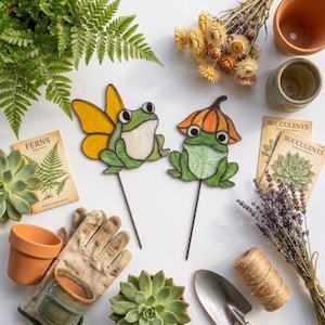 May include: Two stained glass garden stakes featuring green frogs, one with yellow wings and one wearing an orange mushroom cap. The set is surrounded by terracotta pots, gardening gloves, a metal trowel, twine, dried flowers, and seed packets.