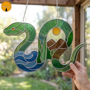 Könnte beinhalten: Glasmalerei-Schlange mit grünem Kopf und Körper, blauem Wasser und einer Landschaft mit gelber Sonne und braunen Bergen. Die Schlange hängt an einer silbernen Kette.