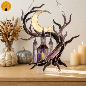 May include: A stained glass suncatcher featuring a purple castle silhouette framed by dark, twisted tree branches and a pale yellow crescent moon. The piece is suspended by a metal chain and rests on a clear acrylic stand, showcasing intricate glass textures.