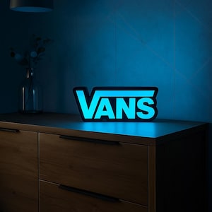 May include: A glowing blue and black "VANS" logo sign. The sign is illuminated and sits on a wooden cabinet. The background is a blue wall with a geometric pattern. A black pendant light hangs above.