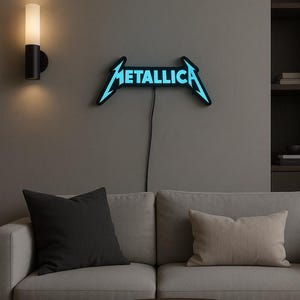 Może przedstawiać: Niebieski neon z napisem "METALLICA" na szarej ścianie. Znak znajduje się nad szarą sofą z czarnymi i beżowymi poduszkami. Widoczny jest również okrągły drewniany stolik kawowy z książką i lampą ścienną.