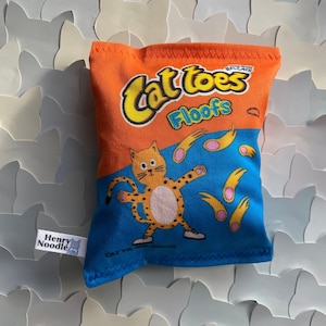 May include: A plush toy shaped like a bag of cat treats. The bag is orange and blue with the text "Cat Toes Floofs" and a cartoon cat with a paw print on its back. The toy is made by Henry Noodle.