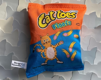 May include: A plush toy shaped like a bag of cat treats. The bag is orange and blue with the text "Cat Toes Floofs" and a cartoon cat with a paw print on its back. The toy is made by Henry Noodle.