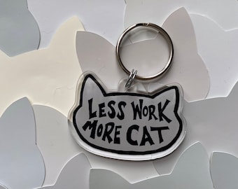May include: A clear acrylic keychain shaped like a cat with the text "Less Work More Cat" in black lettering. The keychain has a silver metal ring.