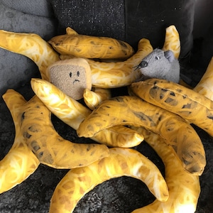 May include: A collection of yellow banana-shaped pillows with a brown, sad-faced sponge pillow in the center. A gray plush mouse pillow is on the right side of the image.