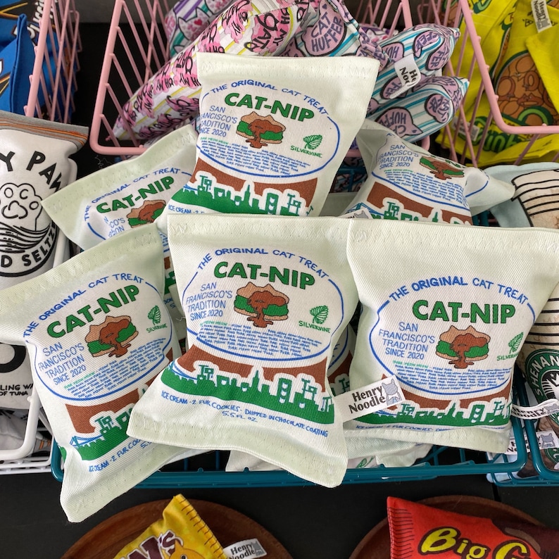 May include: A close-up of several white fabric catnip toys with a brown and blue graphic design. The toys are labeled "The Original Cat Treat Cat-nip" and feature a city skyline, a tree, and a catnip plant. The toys are arranged in a basket.