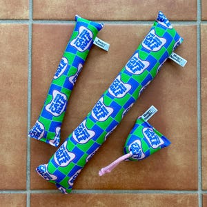 May include: Three fabric pet toys in a blue and green checkered pattern with the text "Dogs Drool Cats Rule" printed in white and pink. Each toy features a small white tag labeled "Henry Noodle" and one toy includes a short pink rope attachment.