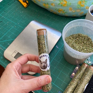 May include: A glass tube filled with dried catnip, labeled "Henry Noodle". The tube has a cork stopper and a sticker with a cat's face on it. There are other tubes of catnip in the background.