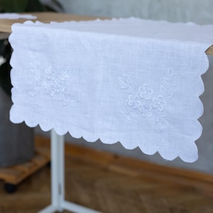 May include: White linen table runner with embroidered floral details along the scalloped edge. The runner is placed on a wooden table, showcasing its delicate design and texture. A decorative piece for home decor.