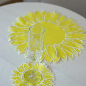 May include: A clear champagne flute rests on a yellow sunflower-shaped coaster, with a larger matching placemat in the background. The coaster and placemat have white outlines, creating a bright, cheerful design. The items are on a white tablecloth.