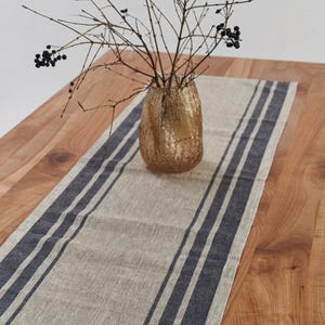 Puede incluir: Camino de mesa de lino beige con tres rayas azul marino, exhibido sobre una mesa de madera. Un jarrón dorado contiene ramas con bayas negras. El camino de mesa añade un toque decorativo.