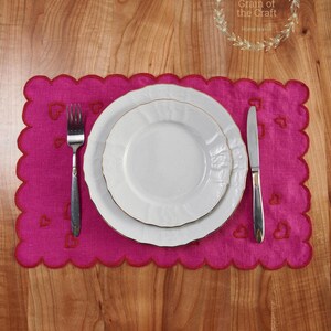 May include: A pink linen placemat with a scalloped edge and embroidered hearts. A white plate with a gold rim is on top of the placemat, along with a silver fork and knife.
