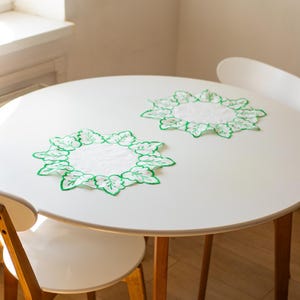 Op de afbeelding: Twee witte en groene placemats in de vorm van bladeren op een witte ronde tafel. De placemats hebben een wit midden met een groene geborduurde bladrand. De tafel wordt omringd door witte stoelen met houten poten.