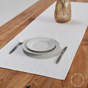 May include: A white linen table runner with a place setting of two plates, a fork, and a knife. The plates have a gold trim. A decorative vase sits at the top of the runner on a wooden table.