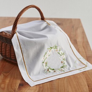May include: A white linen cloth with a gold border and a snowdrop floral embroidery, draped over a brown wicker basket. The embroidery forms a wreath shape. The basket has a woven handle.