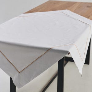 May include: A white tablecloth with a tan border is draped over a wooden table. The tablecloth is square and covers most of the table's surface. The table has black metal legs.