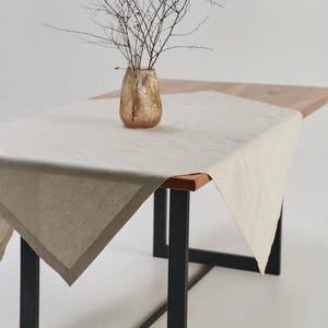 May include: A beige linen tablecloth with a darker border is draped over a wooden table with black metal legs. A gold-colored vase holds dried branches, adding a natural element to the scene. The table is set against a white background.
