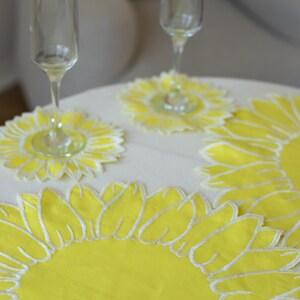May include: A table setting featuring two champagne flutes and yellow sunflower-shaped coasters and placemats. The coasters and placemats have a bright yellow center with white embroidered petal outlines, creating a cheerful, summery vibe.