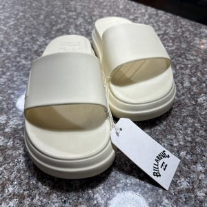 以下が含まれることがあります： オフホワイトのスライドサンダル。厚底で、上部に幅広のストラップ、フットベッドはテクスチャード加工。白いタグには「Billabong」のロゴ。カジュアルな夏の装いに最適なサンダルです。