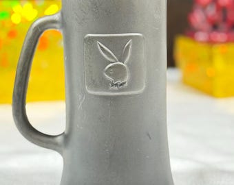 Peut inclure: Un mug en métal gris avec une anse et une base évasée. Le mug présente le logo du lapin Playboy en relief sur le devant. L'intérieur du mug est sombre. L'arrière-plan est flou, avec des touches de jaune et de rouge.