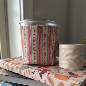 May include: A glass jar wrapped in a fabric sleeve featuring vertical stripes of red, green, and cream with a pattern of candy canes and small red hearts. A small fabric tag labeled Glacier & Pine is attached to the side of the sleeve.