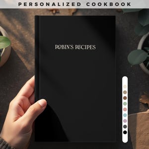 Puede incluir: Un libro de cocina negro con el texto "ROBIN'S RECIPES" en una fuente decorativa. Las palabras "PERSONALIZED COOKBOOK" están en la parte superior. El libro está sostenido por una mano, con plantas en el fondo. Una paleta de colores está a la derecha.