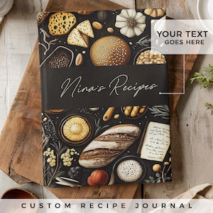 Op de afbeelding: Een zwart, gepersonaliseerd receptenboek met een bloemen- en voedselontwerp. De omslag heeft een handgeschreven titel "Nina's Recipes" en de tekst "YOUR TEXT GOES HERE".