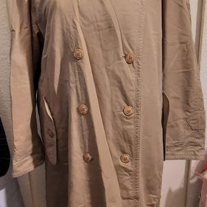May include: A tan trench coat with a double-breasted design. The coat features a classic collar, button closures, and pockets. The coat is displayed on a mannequin.