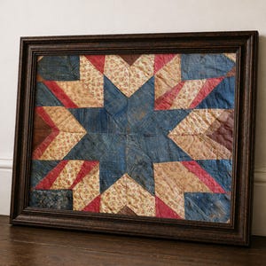 Puede incluir: Una pieza de arte textil enmarcada que presenta un patrón central de estrella de ocho puntas. El bloque de edredón está compuesto por varios parches de tela en tonos azul profundo, rojo descolorido, marrón y crema con un pequeño estampado floral.