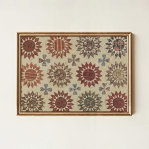 May include: A framed vintage quilt artwork with a repeating pattern of stylized floral designs in red, brown, and blue hues. The artwork is presented in a gold-coloured wooden frame, enhancing its visual appeal. The quilt is set against a neutral backdrop, highlighting the intricate details.